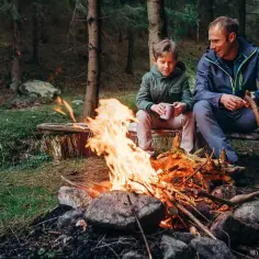 Ojciec i syn siedzą przy ognisku w lesie. Chłopiec trzyma kubek, a mężczyzna patyk. Uważajcie, by nie dostać mandatu za palenie ogniska w lesie!