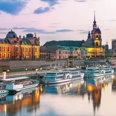 Zachód słońca nad rzeką w Dreźnie. Statki wycieczkowe cumują przy brzegu, odbijając się w spokojnej wodzie. W tle widać oświetlone zabytkowe budowle.