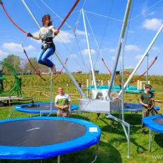 Dziewczyna skacze na eurobungee, unosząc się wysoko nad trampolinami. W tle widać inne atrakcje i ludzi.