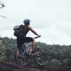 Rowerzysta w kasku i plecaku na skalistym podłożu, patrzący na zalesione wzgórza. Czy to będzie gravel, czy szosa?