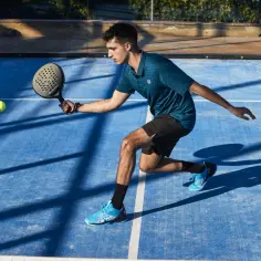 Młody mężczyzna gra w padel, odbijając piłkę rakietą na niebieskim korcie.