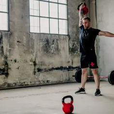 Mężczyzna wykonuje ćwiczenia z kettlebell, podnosząc go nad głowę. W tle sztanga i surowe wnętrze.