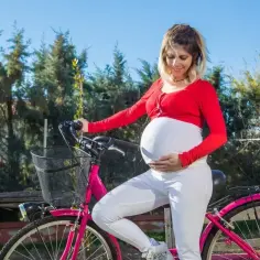 Uśmiechnięta ciężarna kobieta w czerwonej bluzce i białych spodniach opiera się o różowy rower. Czy w ciąży można jeździć na rowerze? Tak, jeśli czujesz się dobrze i zachowasz ostrożność.