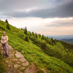 Karpacz-Śnieżka czas wejścia. Kobieta na kamienistej ścieżce, otoczona zielonymi zboczami i lasem, podziwia widok.
