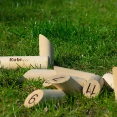 Drewniane kręgle fińskie leżą na trawie. Niektóre są ponumerowane, inne mają logo "Kubi sport".