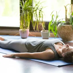 Kobieta w pozycji savasana joga, odpoczywa na macie pośród roślin.