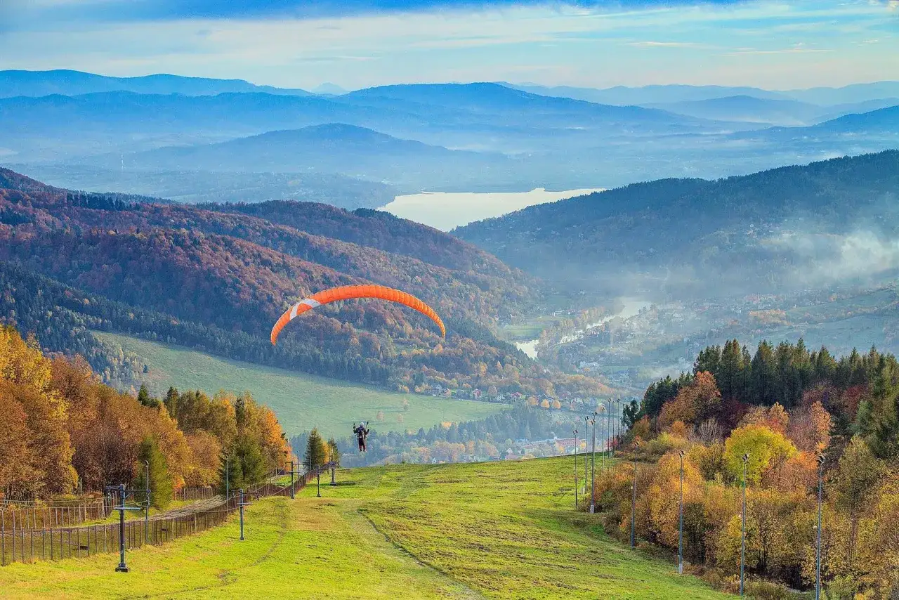 Góra Żar oferuje niesamowite atrakcje. Paralotniarz szybuje nad jesiennym krajobrazem, podziwiając widok na jezioro i dolinę.