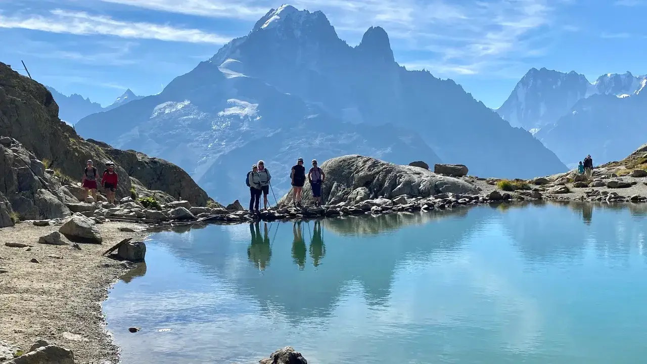 Grupa turystów podziwia majestatyczne szczyty podczas wędrówki Tour du Mont Blanc. Lśniące jezioro odbija niebo i góry.