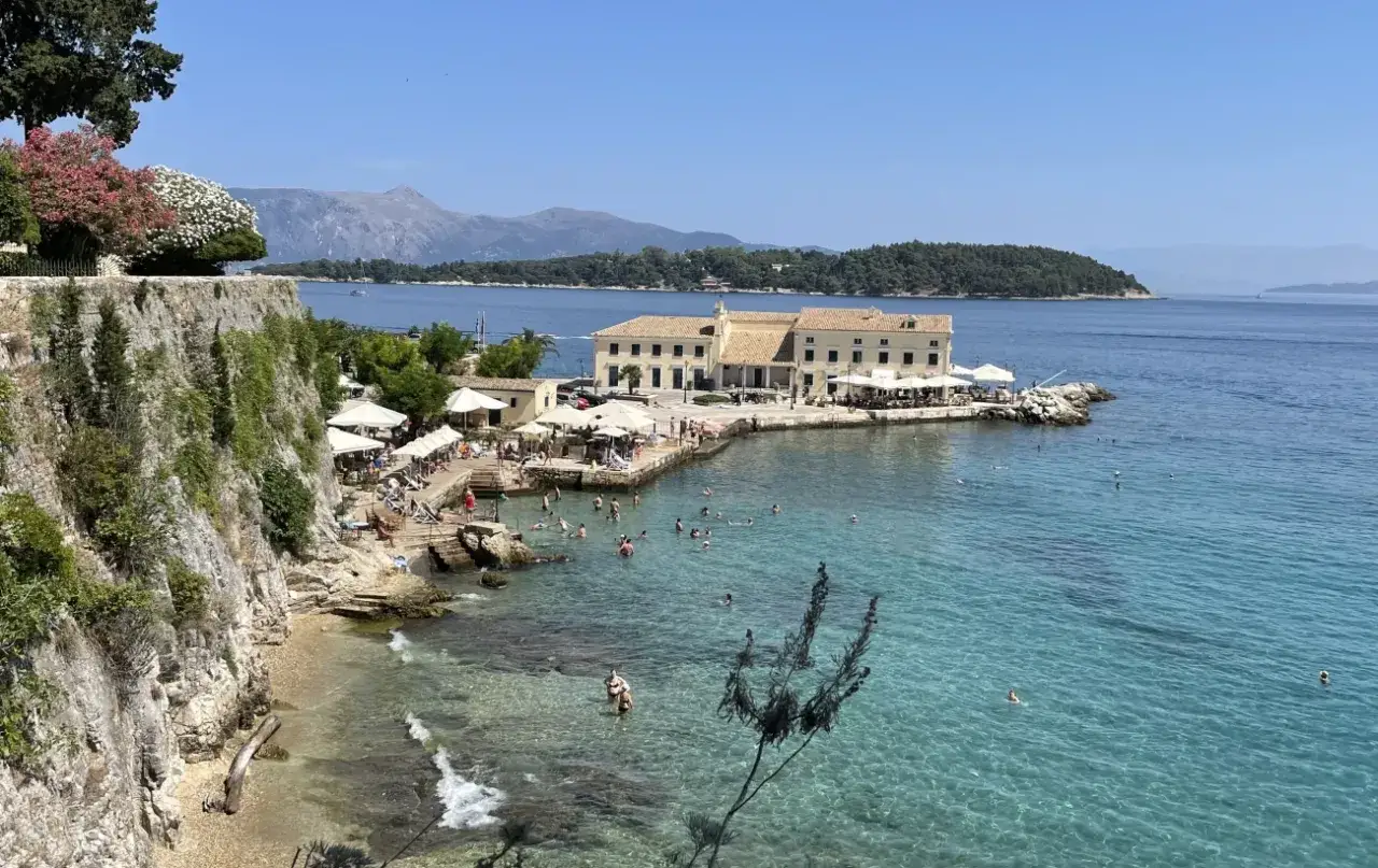 Słoneczna wyspa na Morzu Jońskim z turkusową wodą, kamienistą plażą, budynkiem z restauracją i ludźmi kąpiącymi się w morzu.