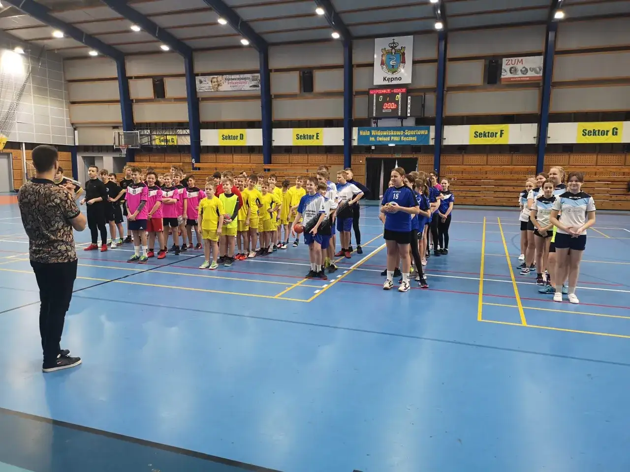 Dzieci w strojach sportowych czekają na rozpoczęcie gry. Trener wyjaśnia zasady, ile trwa mecz piłki ręcznej.