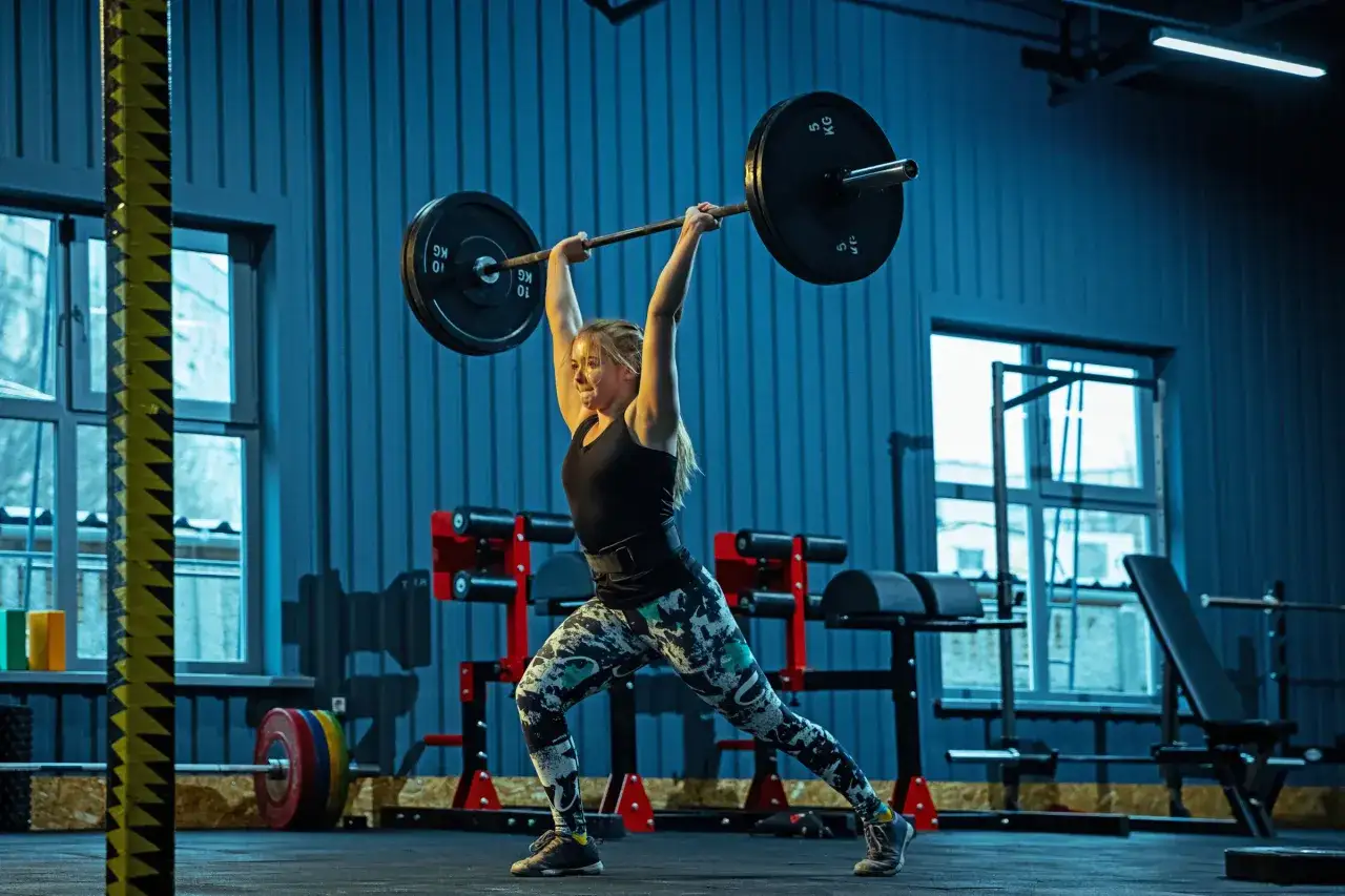 Kobieta w siłowni wykonuje podnoszenie sztangi nad głowę, ćwicząc siłę i technikę, jak w dyscyplinie dwubój olimpijski.