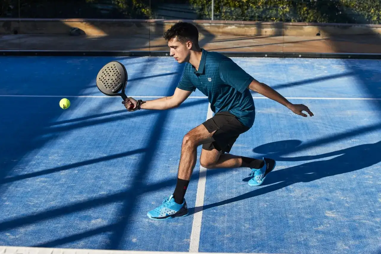 Młody mężczyzna gra w padel, odbijając piłkę rakietą na niebieskim korcie. To świetna zabawa!