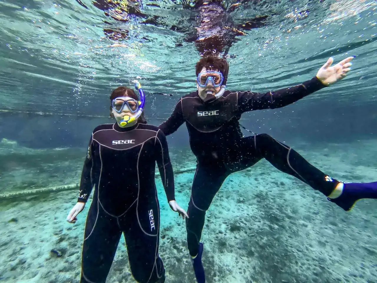 Para pływa pod wodą w kombinezonach, gotowa do snorkelingu. Woda jest przejrzysta, a dno kamieniste.