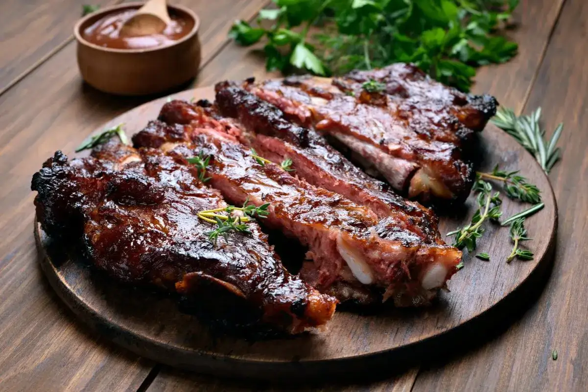 Soczyste, glazurowane żeberka wieprzowe, ozdobione ziołami, podane na drewnianej desce. Obok miseczka z sosem BBQ.