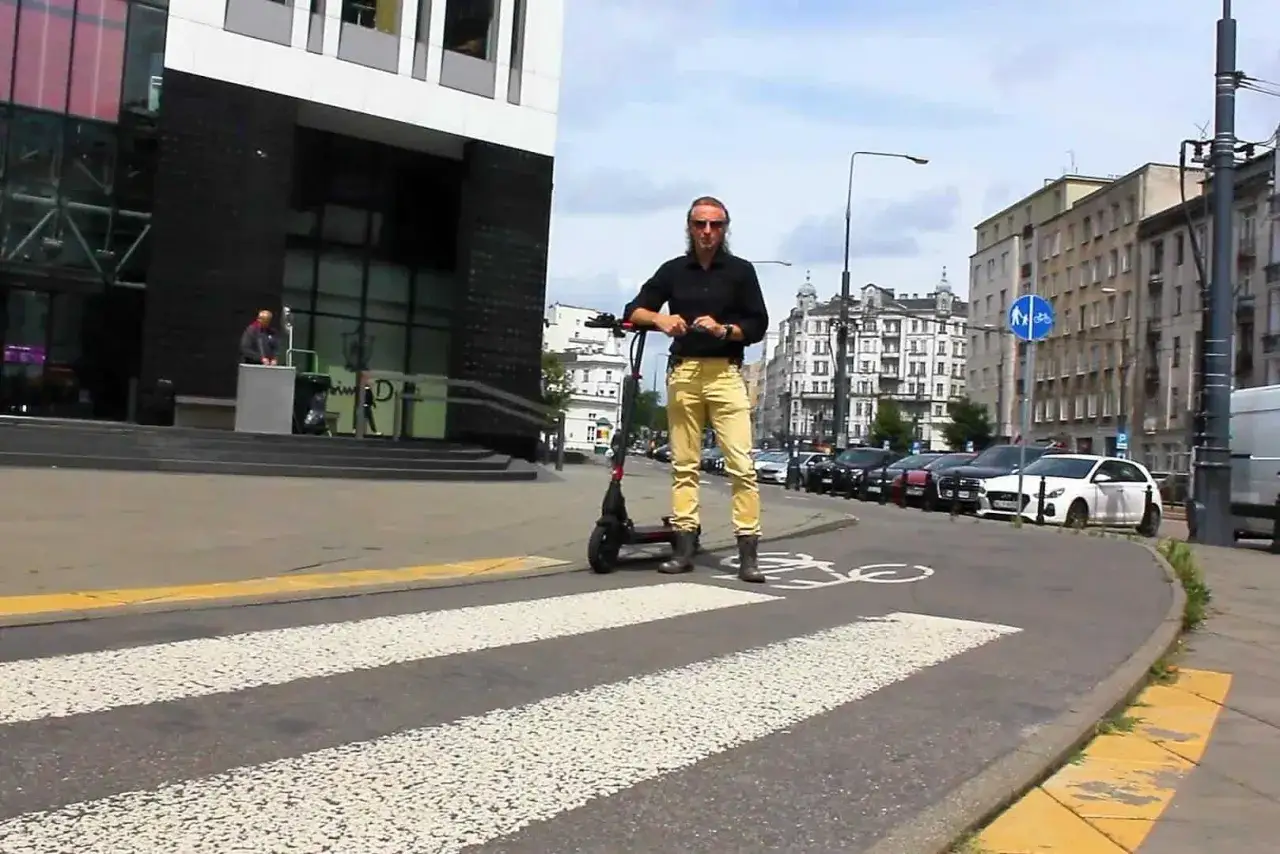 Mężczyzna z hulajnogą elektryczną na pasach. Czy hulajnogą elektryczną można jeździć po chodniku? Znak drogowy sugeruje ścieżkę rowerową.