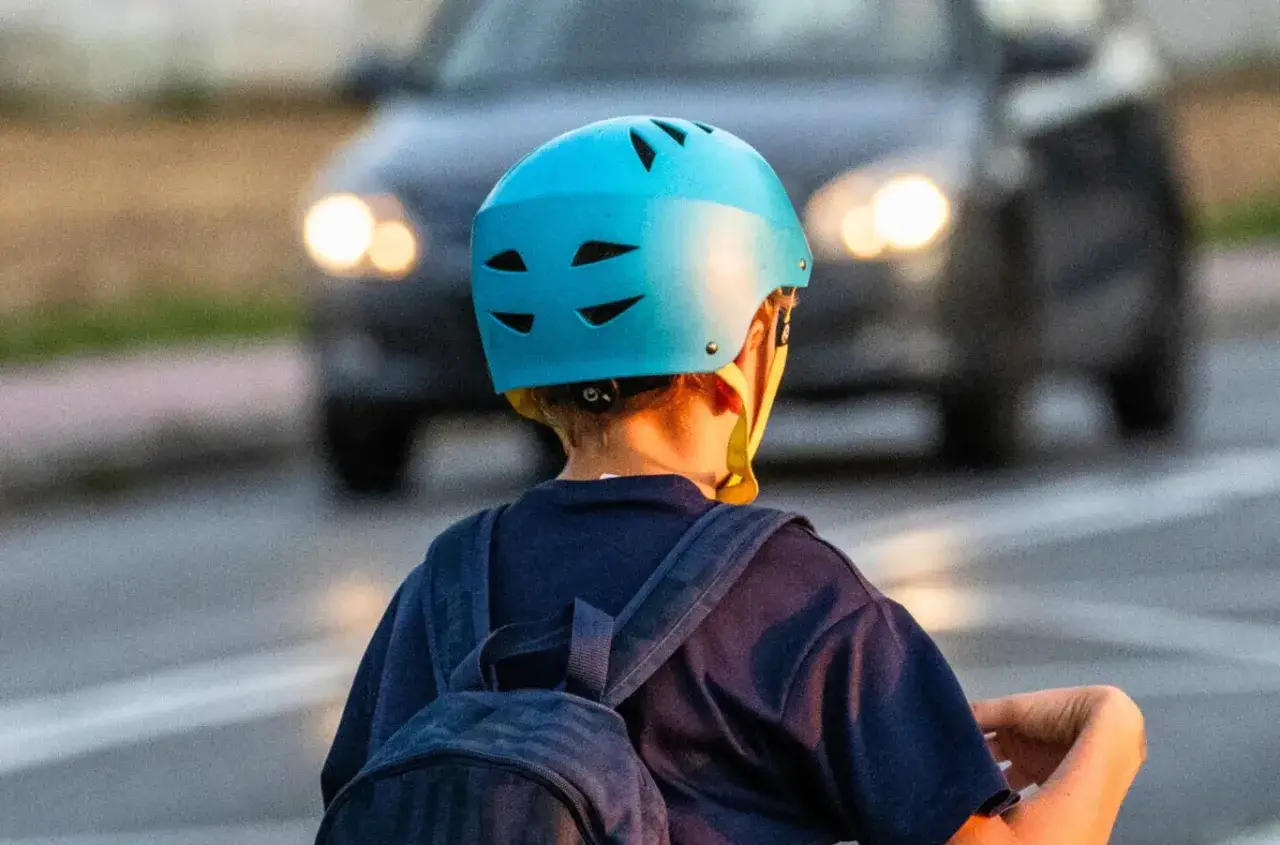 Dziecko w niebieskim kasku na rowerze, z plecakiem, zbliża się do samochodu. Czy kask na rower jest obowiązkowy? Bezpieczeństwo przede wszystkim!