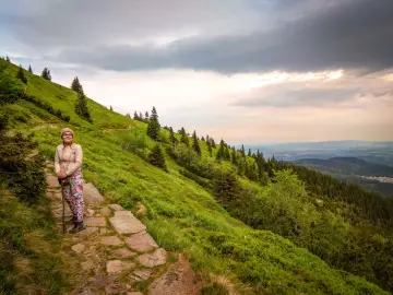 Karpacz-Śnieżka czas wejścia. Kobieta na kamienistej ścieżce, otoczona zielonymi zboczami i lasem, podziwia widok.