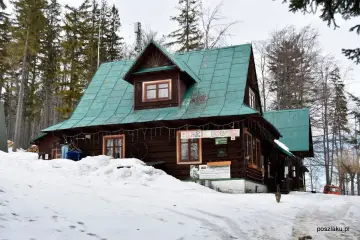 Drewniany domek na przełęczy Przegibek, otoczony ośnieżonym lasem. Na dachu zielona blacha, a na ścianach ozdoby.